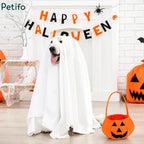 Cute dog wearing a white ghost sheet costume sitting indoors with a Happy Halloween banner, carved pumpkins, and festive holiday decor.