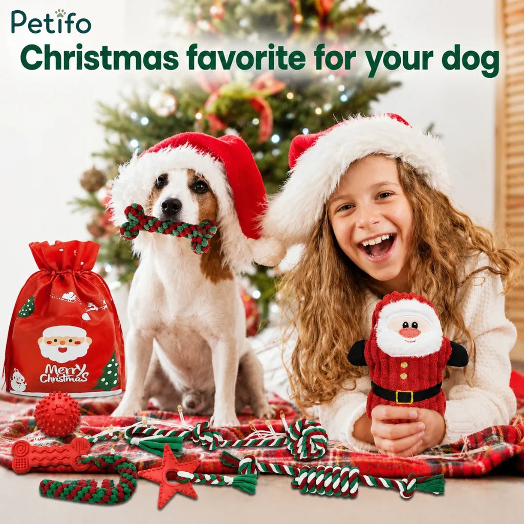 A Christmas scene showing a dog wearing a Santa hat holding a bone toy in its mouth next to a smiling young girl holding a plush Santa toy, with a Christmas tree and a red gift bag in the background.