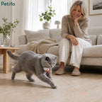 Woman sitting on a sofa smiling as a grey cat enthusiastically chases the red laser dot from its Petifo collar on a wooden floor.